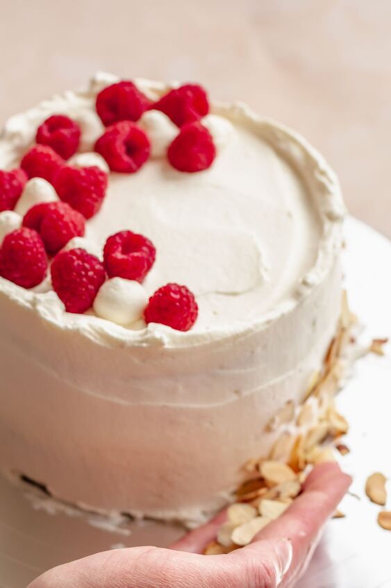 raspberry and almond cake, A hand presses sliced almonds onto the base of the cake and raspberries are on top