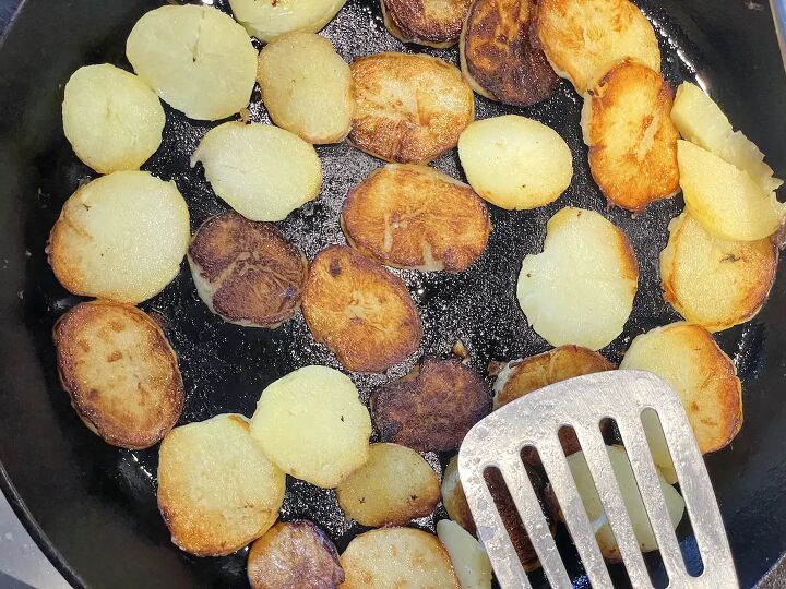 easy german fried potatoes bratkartoffeln, fried potato slices in cast iron skillet with stainless steel spatula