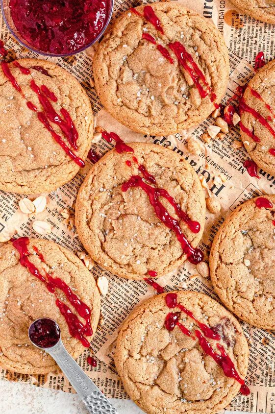 peanut butter and jelly cookies gluten free dairy free, Light golden brown peanut butter cookies topped with white coarse sea salt They are surrounded by chopped peanuts and sitting on a piece of brown newspaper The cookies have been drizzled with bright red strawberry jelly on top