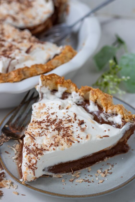 homemade chocolate coconut cream pie, A small pie plate with a slice of chocolate coconut cream pie There s a fork next to the plate and a white pie plate with coconut pie in the background The pie slice is topped with whipped cream toasted coconut and shaved chocolate pieces