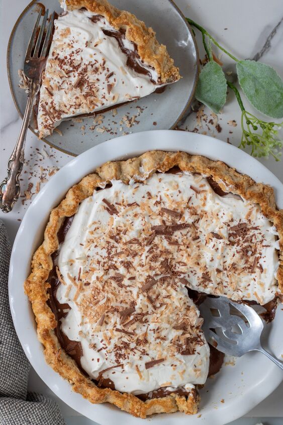 homemade chocolate coconut cream pie, A white pie plate filled with chocolate coconut cream pie There s toasted coconut and chocolate shavings on top There s a small plate in the background with a slice of pie on it There s a green flower in the background