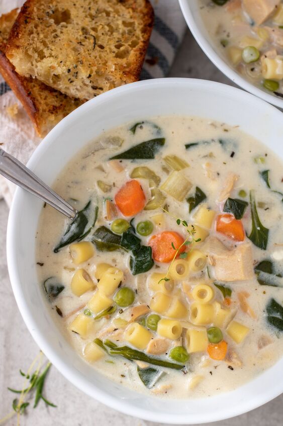 instant pot creamy chicken soup, A white bowl with creamy chicken soup It s got carrots spinach small pasta and peas in it There s garlic bread next to the bowl in the background