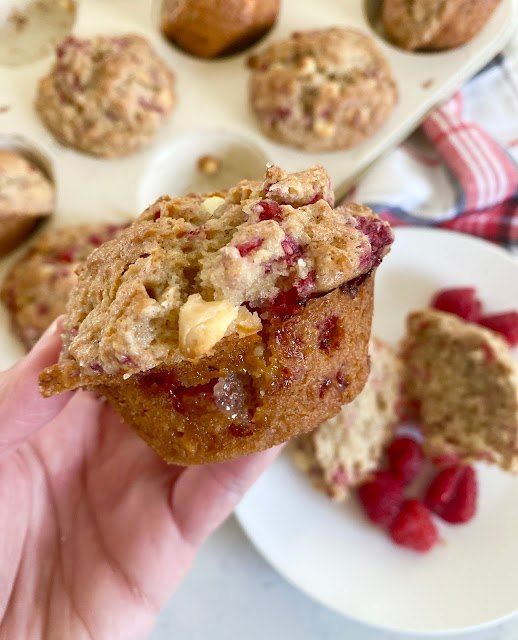 white chocolate raspberry bakery muffins, A hand holding a fresh raspberry muffin with a full muffin tin in the background