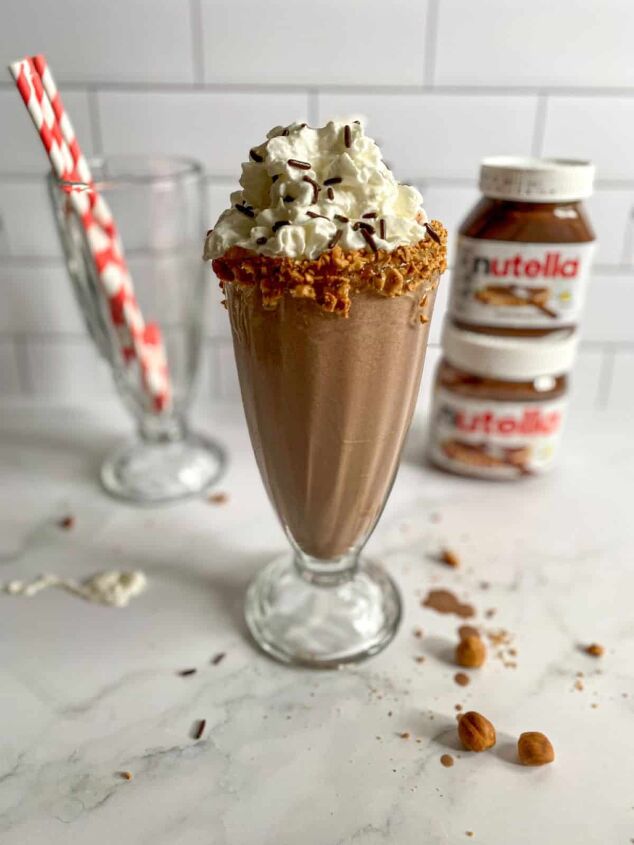 nutella milkshake recipe 3 ingredients, Nutella milkshake in a clear glass topped with whipped cream and sprinkles in a tall old fashioned milkshake glass Jars of Nutella and red straws are in the background