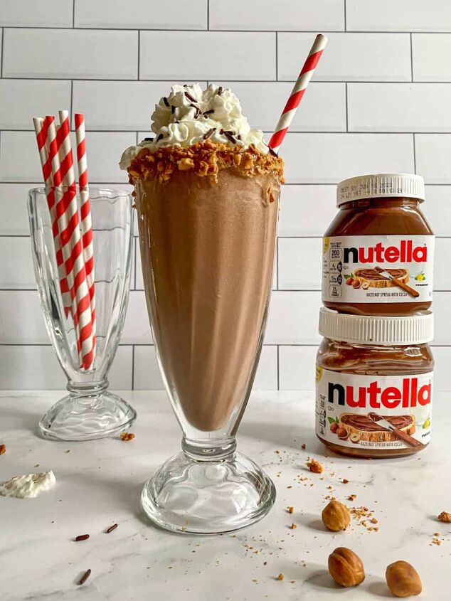 nutella milkshake recipe 3 ingredients, Nutella milkshake topped with whipped cream and sprinkles in a tall old fashioned milkshake glass with a red straw Jars of Nutella and red straws are in the background
