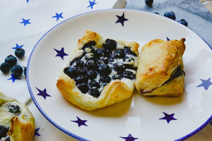easy blueberry puff pastry turnovers, puff pastry blueberry turnovers sitting on a blue and white star plate with raw blueberries in the background on a marble backdrop