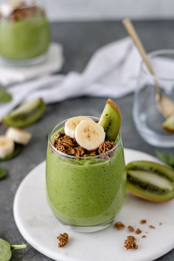 kiwi avocado green smoothie, A full glass of green smoothie sitting on a marble trivet topped with granola banana slices and a kiwi wedge