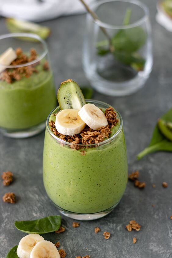 kiwi avocado green smoothie, A glass of a Kiwi Avocado Green Smoothie and a glass filled with some spinach and a spoon are surrounded by granola bits banana slices spinach leaves and kiwi wedges