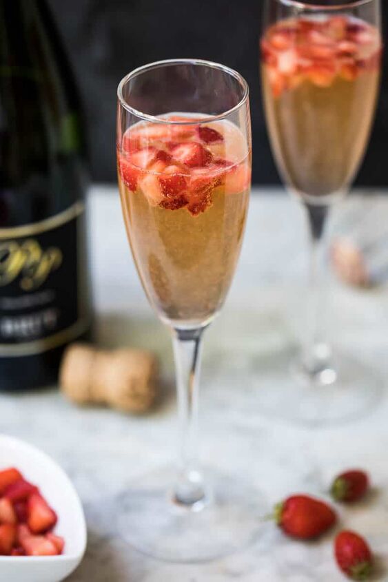 strawberry mimosa, strawberry mimosa in champagne glass with bowl of chopped strawberries a few whole strawberries another glass and champagne bottle in background