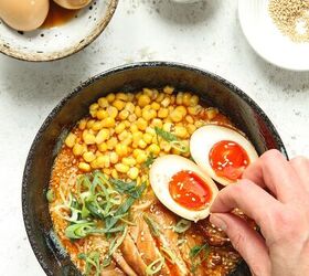 easy spicy chicken miso ramen, Hand adding sesame seeds to a bowl of miso ramen on a white background with three additional bowls containing ramen eggs sesame seeds and sliced scallions