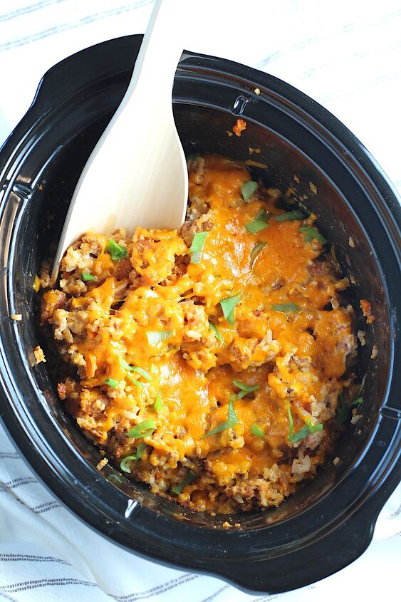 ground pork slow cooker casserole, Crockpot insert on counter with Ground Pork Slow Cooker Casserole inside and melted cheese on top with scallions garnish and a wood spoon in it