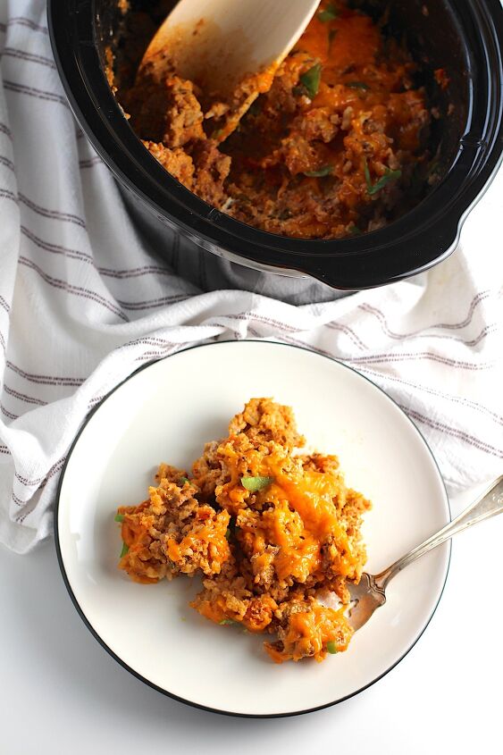 ground pork slow cooker casserole, Plate on counter with Ground Pork Slow Cooker Casserole on it and melted cheese on top A fork is on plate with a bite of the casserole scooped In the background is the slow cooker insert with wood spoon