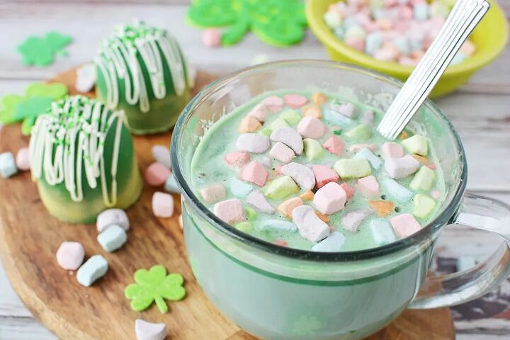 glittery green hot chocolate bombs recipe, Mug of green hot chocolate with rainbow marshmallows sitting on a board next to green hot chocolate bombs and more marshmallows
