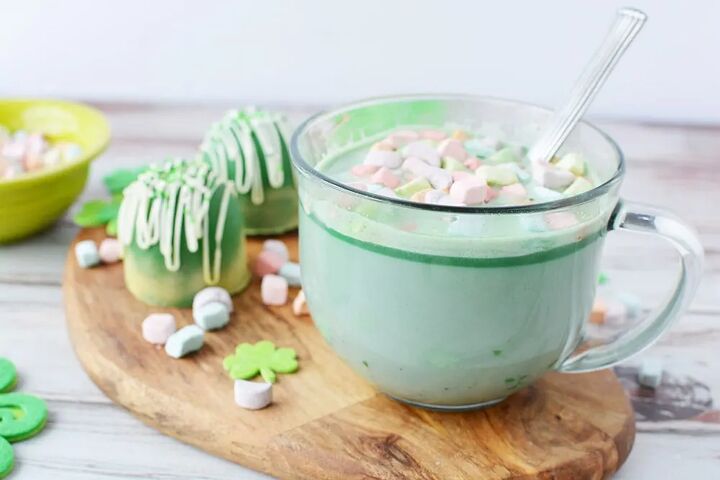 glittery green hot chocolate bombs recipe, Clear mug with green hot cocoa topped with rainbow marshmallows sitting on a board next to hot cocoa bombs