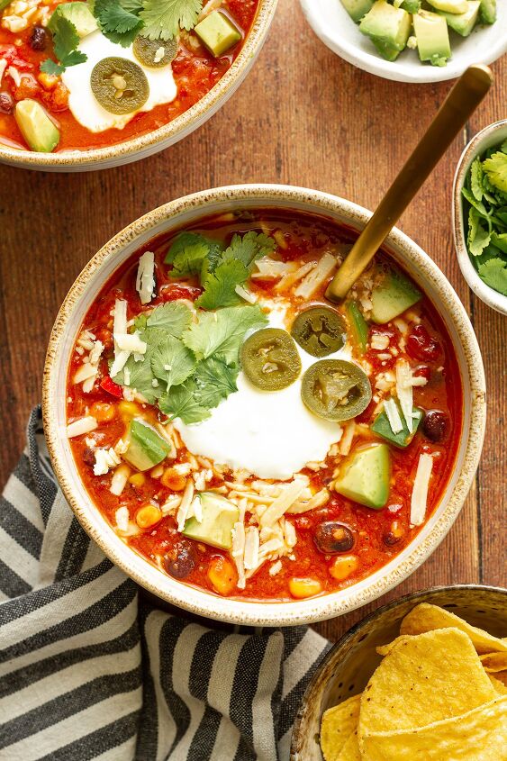 easy spicy mexican chilli bean soup with corn, A bowl of Mexican Bean Soup on a wooden table background with Jalapeno cilantro cheese and avocado toppings and side dishes of chopped avocado cilantro and tortilla chips