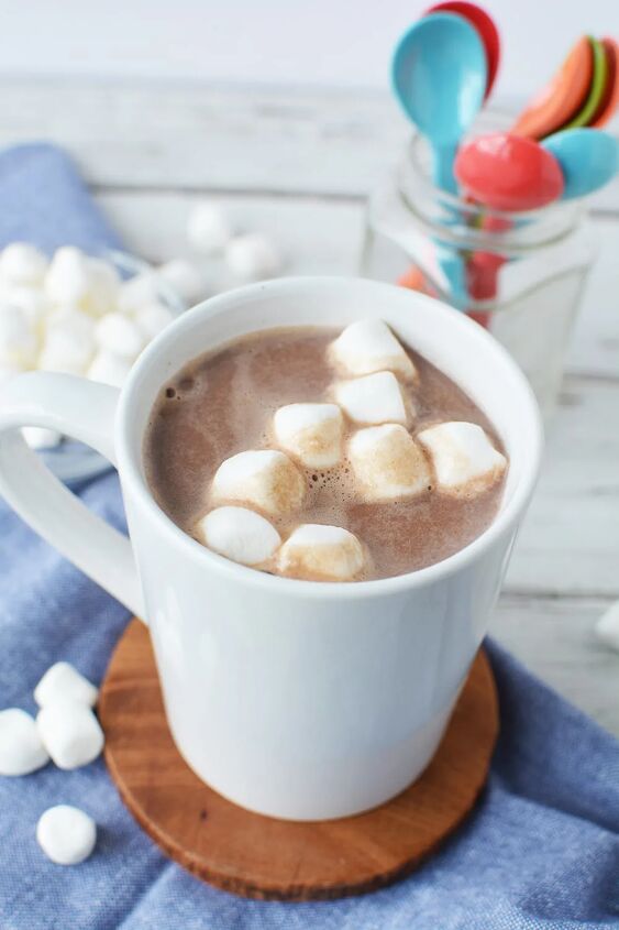extra creamy homemade hot chocolate recipe, Mug of hot cocoa topped with marshmallows on a table with colored spoons in a jar