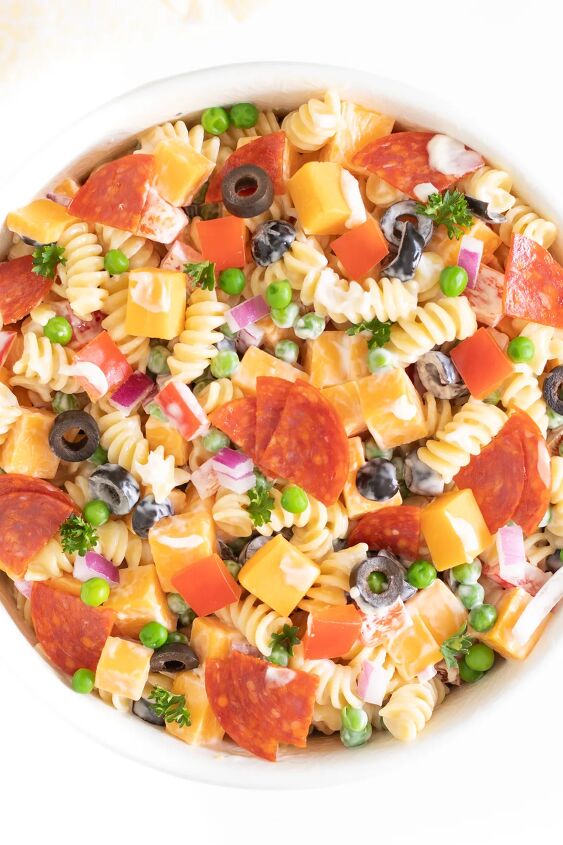 the throwback creamy pasta salad everyone will rave about, up close of pasta salad in a bowl topped with cheese chunks pepperoni quarters parsley chopped red peppers and more