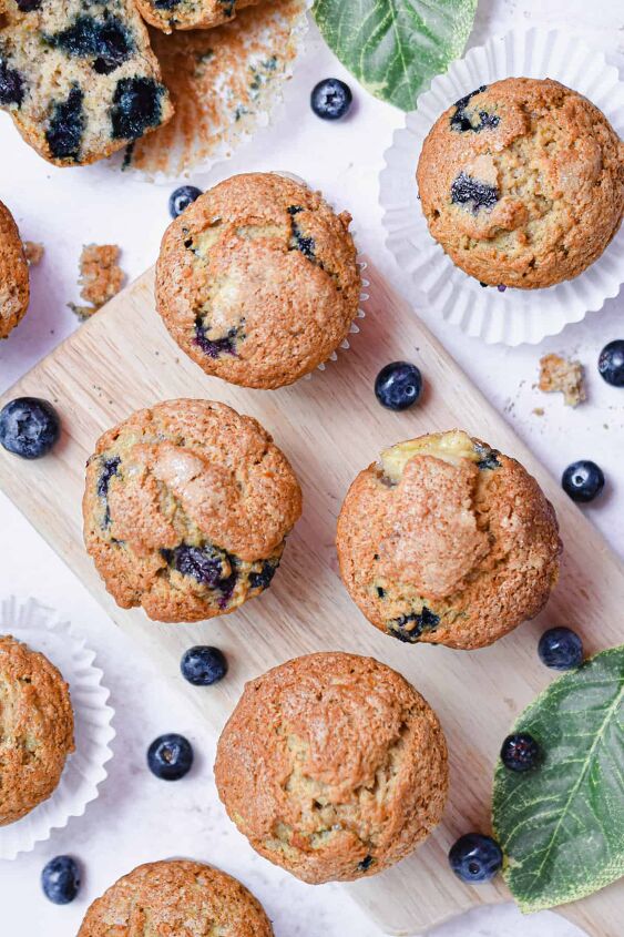 the best banana blueberry muffins so moist, A group of banana blueberry muffins are on a cutting board with leaves and blueberries around them