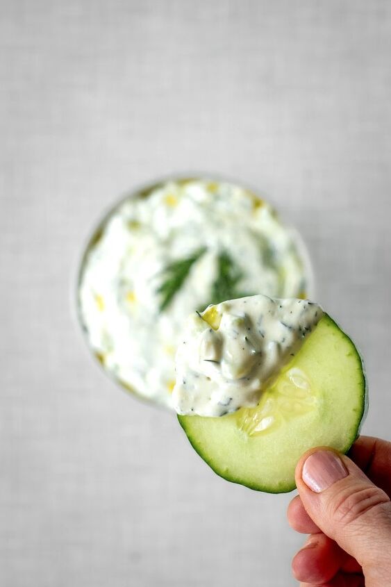 tzatziki, A hand holds a slice of cucumber dipped in tzatziki In the background a bowl filled with tzatziki garnished with dill seen from above