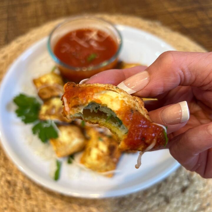 easy air fryer toasted ravioli, A hand holding a bitten air fried toasted ravioli over a plate of air fryer raviolis with marinara sauce