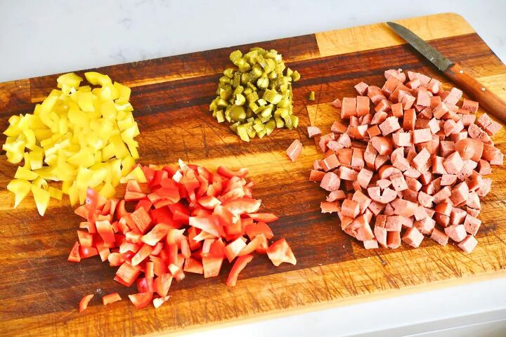 classic creamy german pasta salad nudelsalat, wooden chopping board with chopped yellow bell peppers red bell peppers cornichons and hot dogs