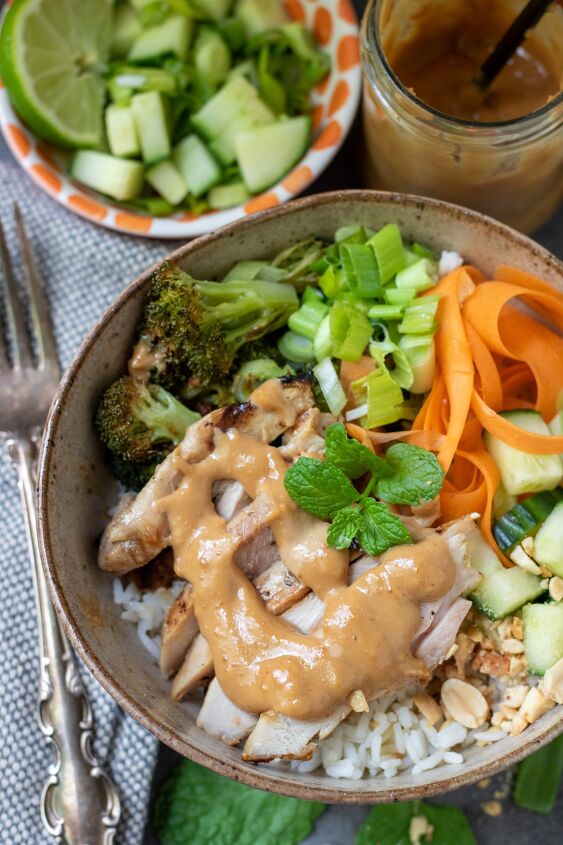 thai chicken rice bowls with peanut sauce, A bowl filled with rice roasted broccoli carrots and diced cucumber There s sliced grilled chicken and a peanut sauce drizzled on top There s a fork next to the bowl and a small bowl filled with diced cucumber in the background