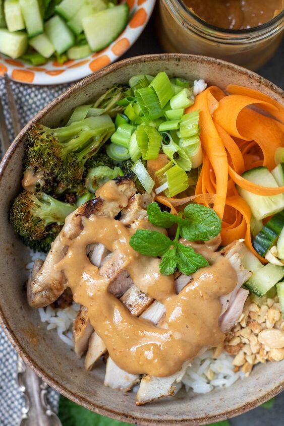thai chicken rice bowls with peanut sauce, A close up of a Thai chicken rice bowl There s a peanut sauce drizzled on top The bowl has ribboned carrots diced cucumbers scallions and broccoli There s a sprig of fresh mint on top of the rice bowl