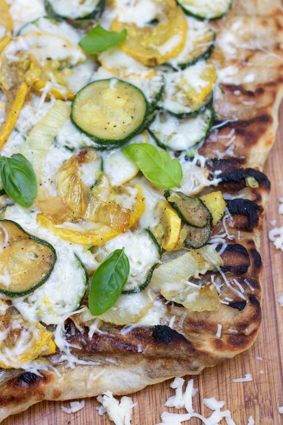 easy summer squash grilled pizza, Grilled pizza on a wooden cutting board with shredded cheese sprinkled on the board The crust has slight char marks on it and it s topped with summer squash onions and fresh basil