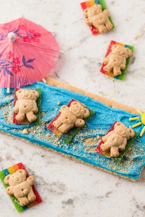 how to make beach day graham crackers, angled down photo of beach day graham crackers with teddy grahams laying on rainbow candy drink umbrella nearby