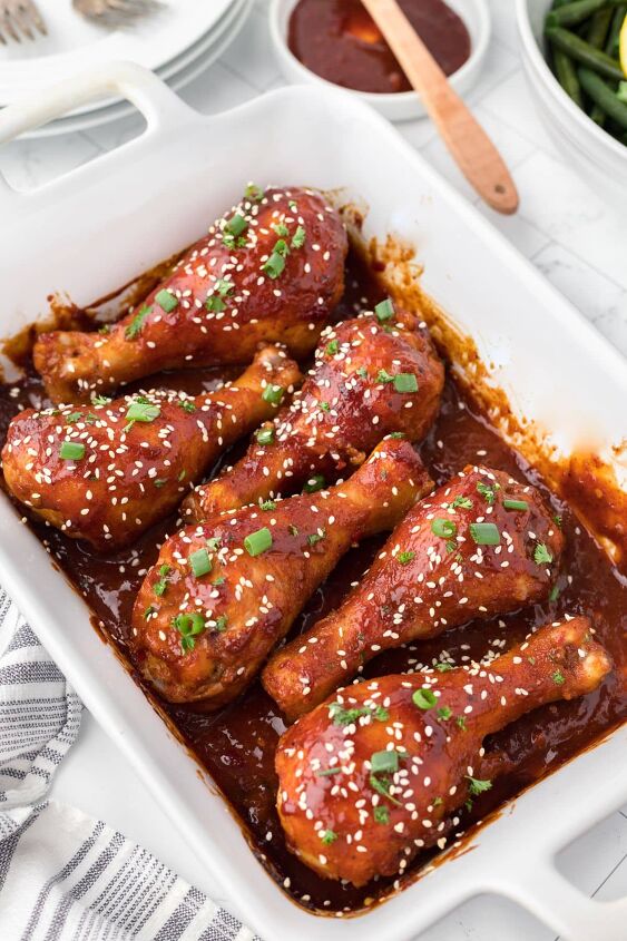 baked bbq chicken drumsticks, BBQ drumsticks in a white baking dish with a blue striped linen on the side and bbq sauce in a small bowl with a brush