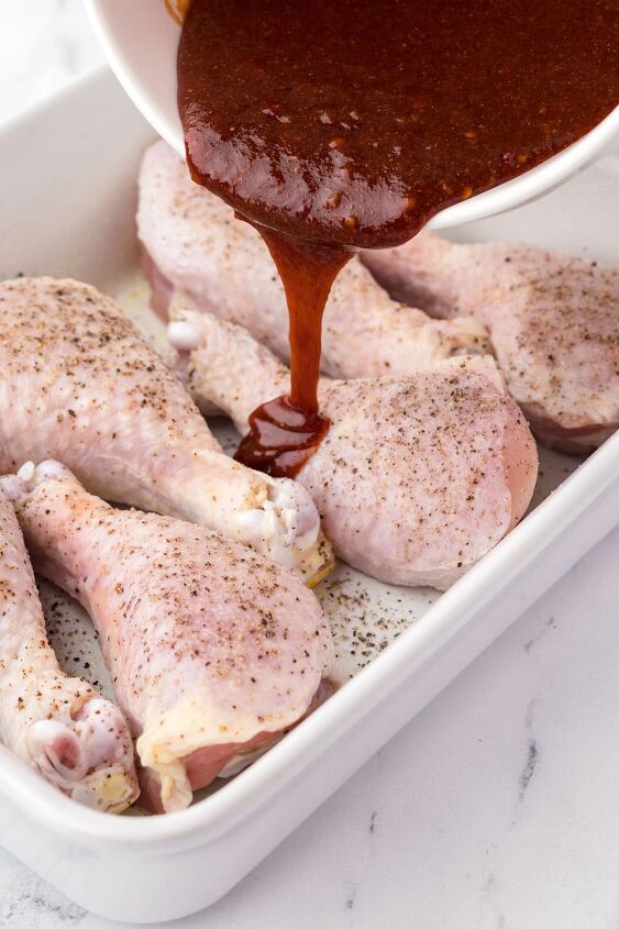 baked bbq chicken drumsticks, raw seasoned chicken drumsticks in a white casserole dish with bbq sauce being poured over them
