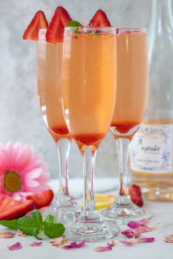 sparkling strawberry rose cocktail, 3 champagne flutes filled with strawberry mint syrup and topped with sparkling ros champagne There s a bottle of the ros in the background with a pink daisy flower and fresh strawberries