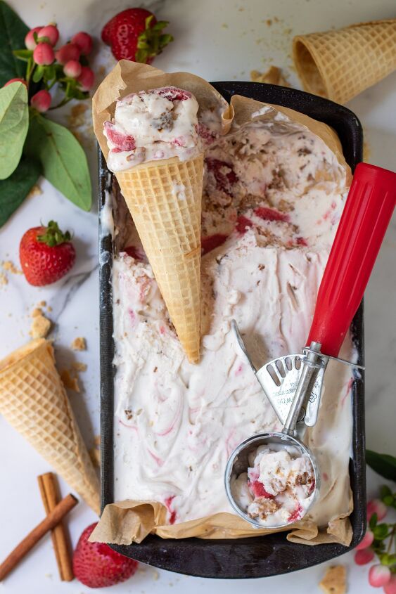 A loaf pan filled with strawberry ice cream There s a red ice cream scoop and two cones of ice cream There s whole strawberries in the background