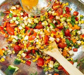 Lime vinaigrette is poured over corn and diced vegetables in a stainless steel bowl with a wooden spoon