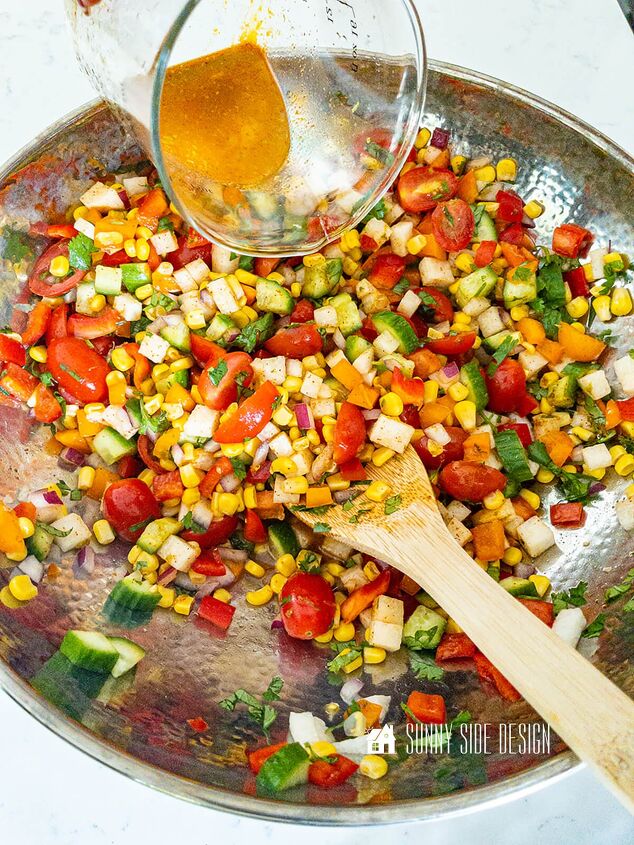 Lime vinaigrette is poured over corn and diced vegetables in a stainless steel bowl with a wooden spoon