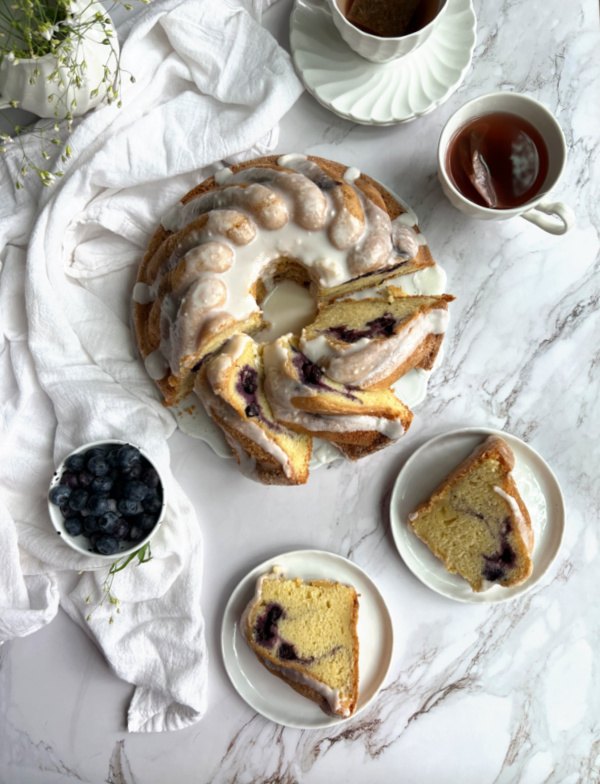 bonne maman blueberry swirl bundt cake, blueberry swirl bundt cake cut on a platter with 2 pieces on white plates and a cup of tea and extra blueberries nearby