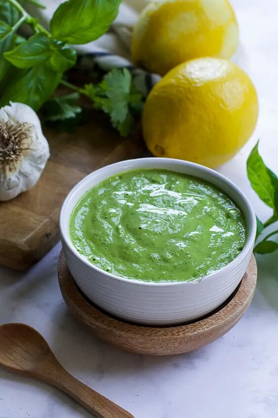 mediterranean herbed tahini sauce, a bowl with green sauce with a small wooden spoon on a table next to it a lemon herbs and a head of garlic on a cutting board