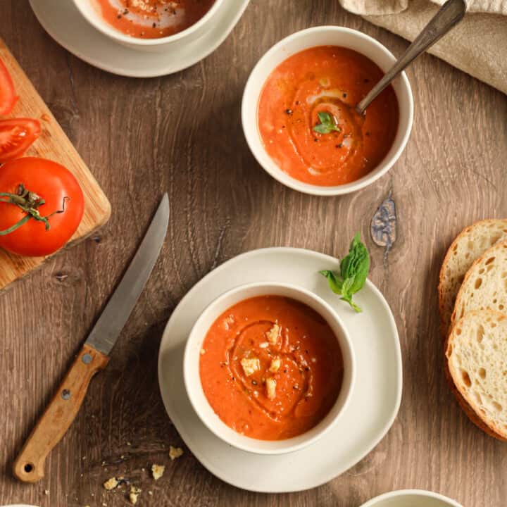 cold lemon capellini salad with arugula angel hair pasta, Gluten free tomato soup in three white bowls on a wood surface with a knife and tomato on a cutting board next to it and bread as well as a napkin