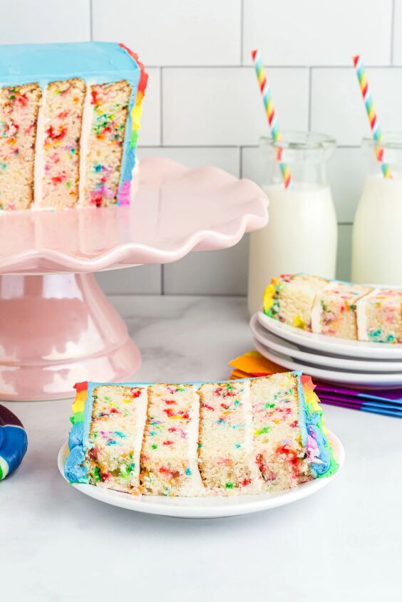 how to make a funfetti rainbow cake perfect for every celebration, pretty up close slice of rainbow cake on a small white dish