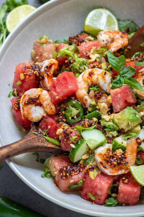 spicy watermelon salad with shrimp, A large cream colored serving bowl filled with spicy watermelon salad There s a wooden serving spoon in the salad It s topped with chili crunch oil and grilled shrimp