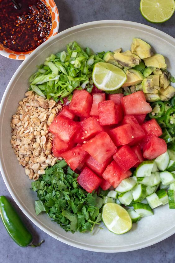 spicy watermelon salad with shrimp, A large round serving bowl with cubes of watermelon diced cucumbers diced avocado sliced jalape os sliced scallions crushed peanuts and fresh chopped herbs There s a small bowl filled with chili crunch oil in the background