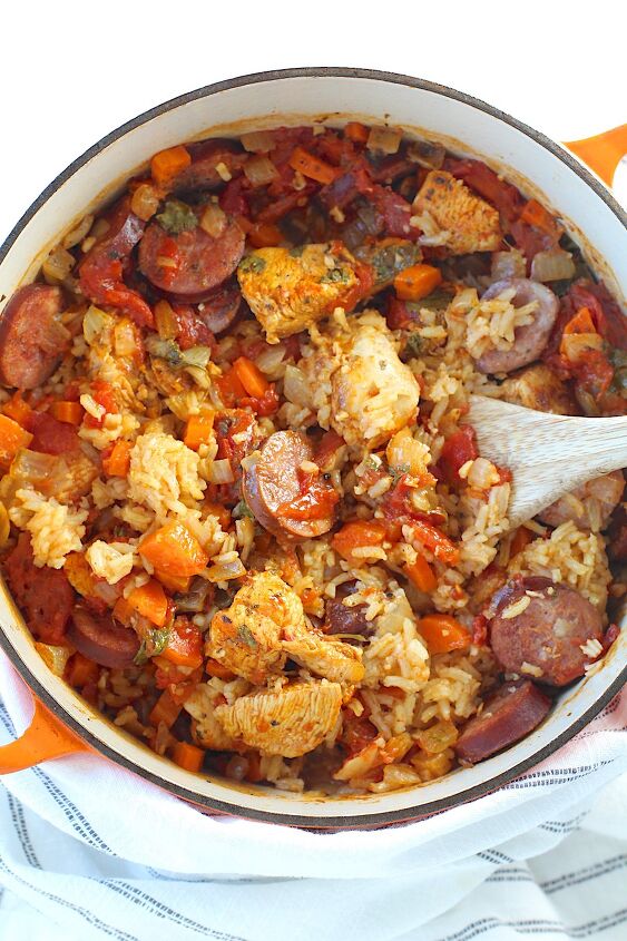 galinhada dutch oven chicken and rice, Galinhada Dutch Oven Chicken and Rice with slices of sausage carrots onions and more in a dutch oven with wood spatula on counter with blue and white towel