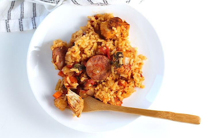 galinhada dutch oven chicken and rice, Galinhada Dutch Oven Chicken and Rice with slices of sausage carrots onions and more on a plate with bamboo work holding a bite of chicken