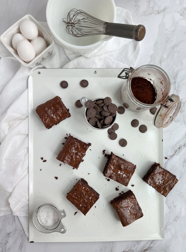condensed milk brownies, sweetened condensed milk brownies on a white pan with a basket of eggs a white bowl and whisk and a jar of cocoa powder
