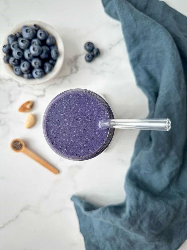 blueberry protein shake, Looking down at a marble table with a blueberry smoothie measuring spoon with chia seeds and nuts A blue kitchen towel is next to the glass