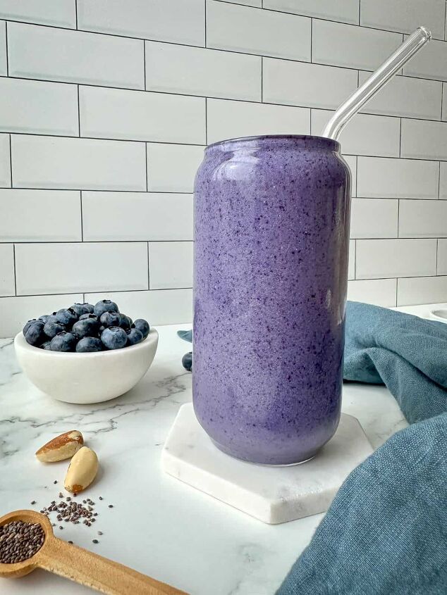 blueberry protein shake, A purple smoothie in a clear glass A bowl of blueberries Brazil nuts and chia seeds are on a marble table with a blue kitchen towel
