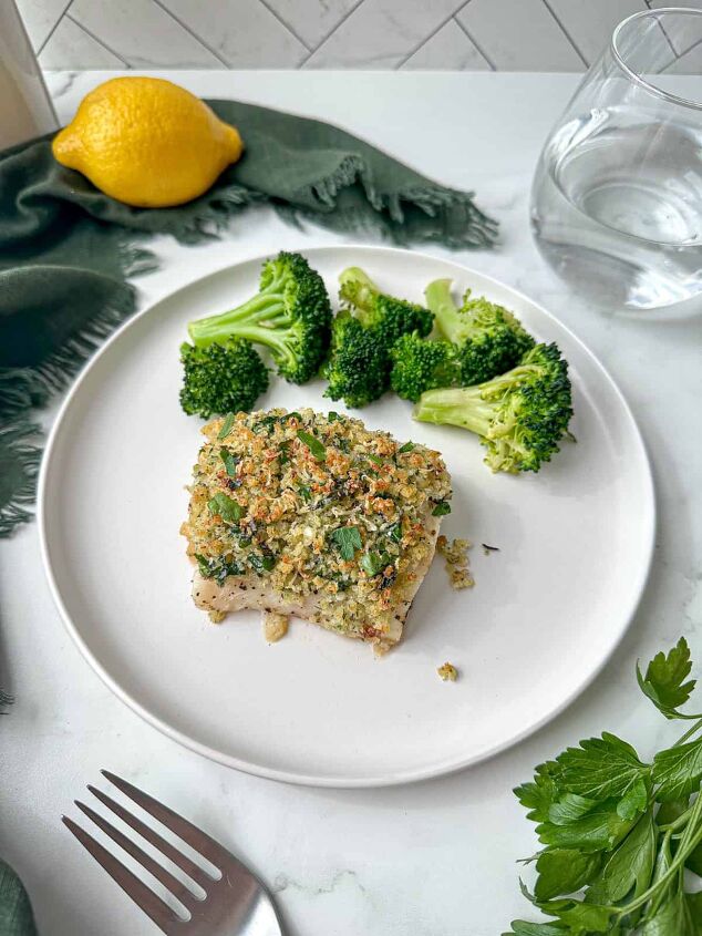 baked panko crusted cod, Parmesan and panko crusted cod on a white plate with a side of broccoli A green towel lemon and glass of water are next to the plate