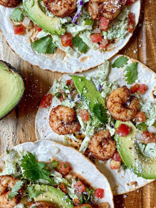 baked panko crusted cod, Three air fried shrimp tacos with slaw avocado cilantro and creamy jalapeno salsa are open face on a cutting board Lime wedges and an avocado are on the side