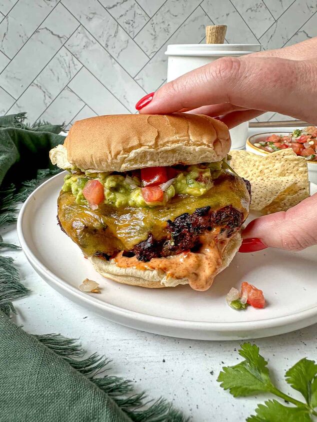 southwest burger with chipotle mayo, A hand with red nails is grabbing a taco burger topped with guacamole pico de gallo and spicy mayo dipping out from the bottom bun