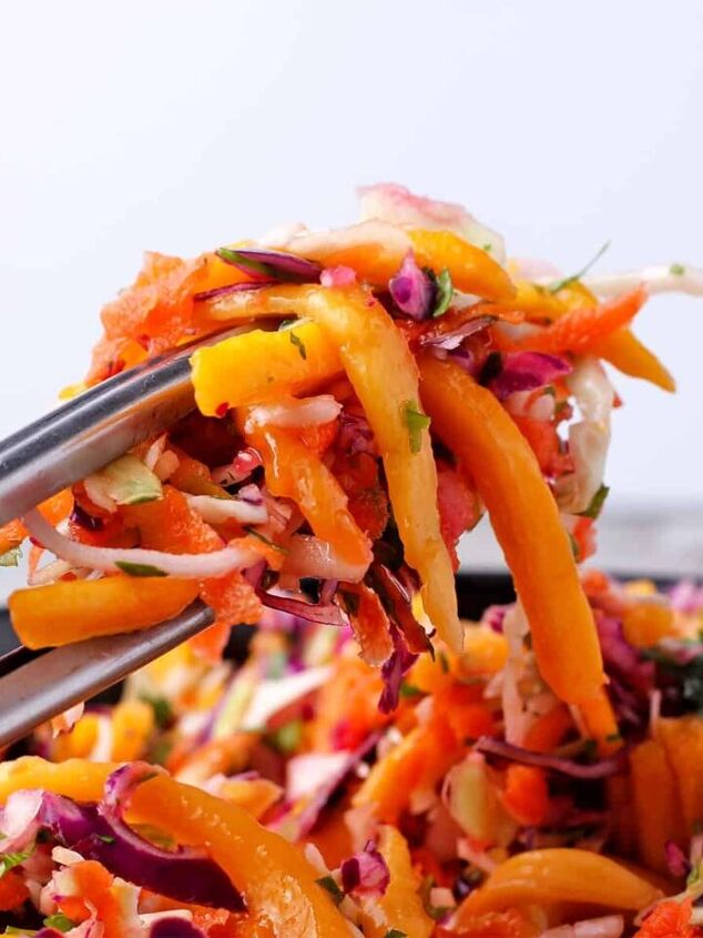 lentil pomegranate salad with tahini dressing, Tongs hold coleslaw with green and red cabbage carrots chopped cilantro and mango strips over a bowl of slaw